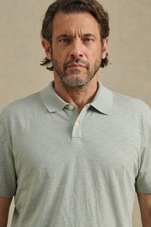 Man with short brown hair and a beard wears the MORRIS POLO by Velvet by Graham & Spencer, a light gray cotton slub polo with a ribbed collar and short sleeves, standing against a plain beige background.