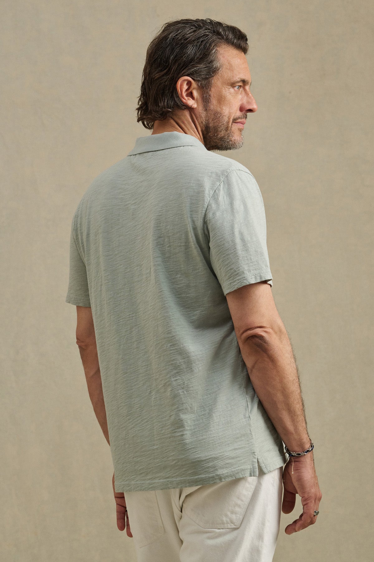 A man with short hair and a beard stands sideways, facing left, wearing the MORRIS POLO by Velvet by Graham & Spencer—a light grey cotton slub polo with a ribbed collar and short sleeves—paired with light-colored pants against a neutral background.