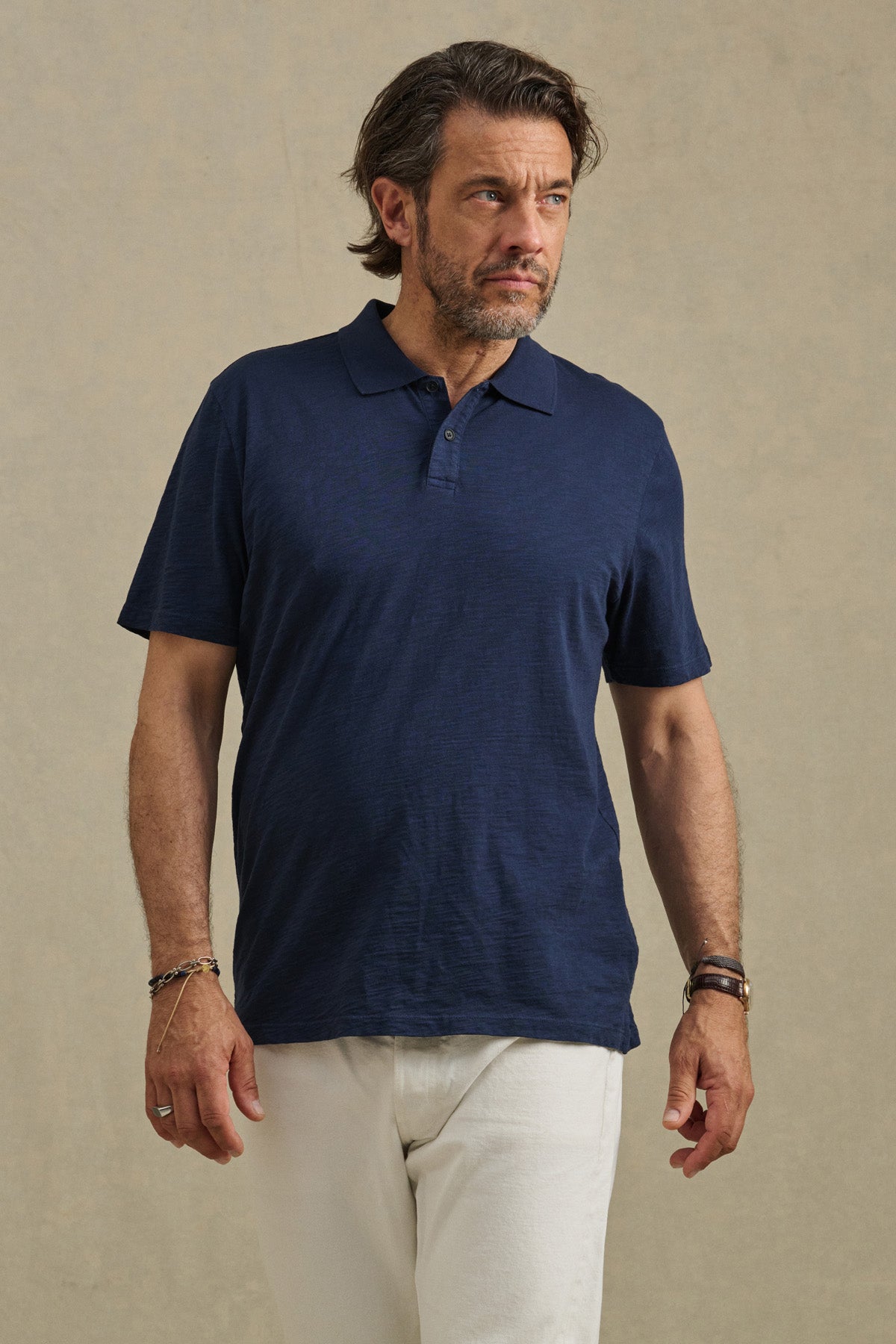 A man with medium-length brown hair and a beard wears the MORRIS POLO by Velvet by Graham & Spencer in navy blue with white pants, standing against a plain beige background.