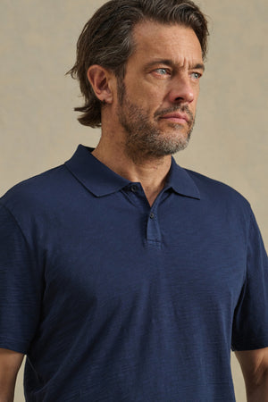 A middle-aged man with brown hair and a beard stands against a plain beige background, wearing the navy blue MORRIS POLO by Velvet by Graham & Spencer, featuring cotton slub fabric and a two-button placket.