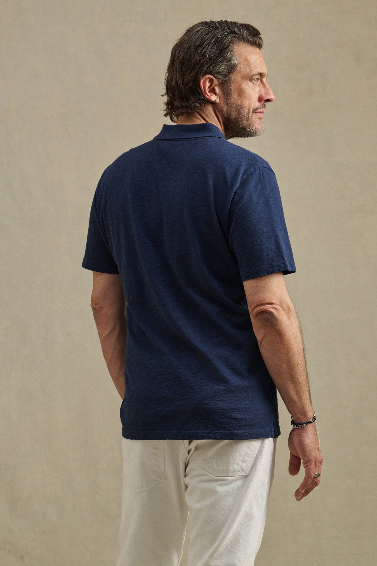 A man stands facing away, wearing Velvet by Graham & Spencer's MORRIS POLO in navy blue cotton slub with a ribbed collar and two-button placket, paired with white pants against a beige background.-41374410473665