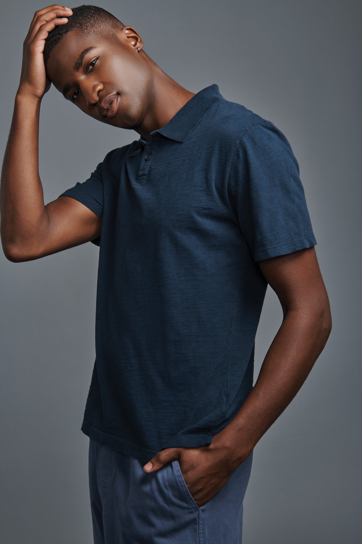 A man wearing the MORRIS POLO by Velvet by Graham & Spencer—featuring navy blue cotton slub fabric, a ribbed collar, and short sleeves—paired with blue pants, stands against a gray background with one hand touching his head and the other in his pocket.-41776983507137
