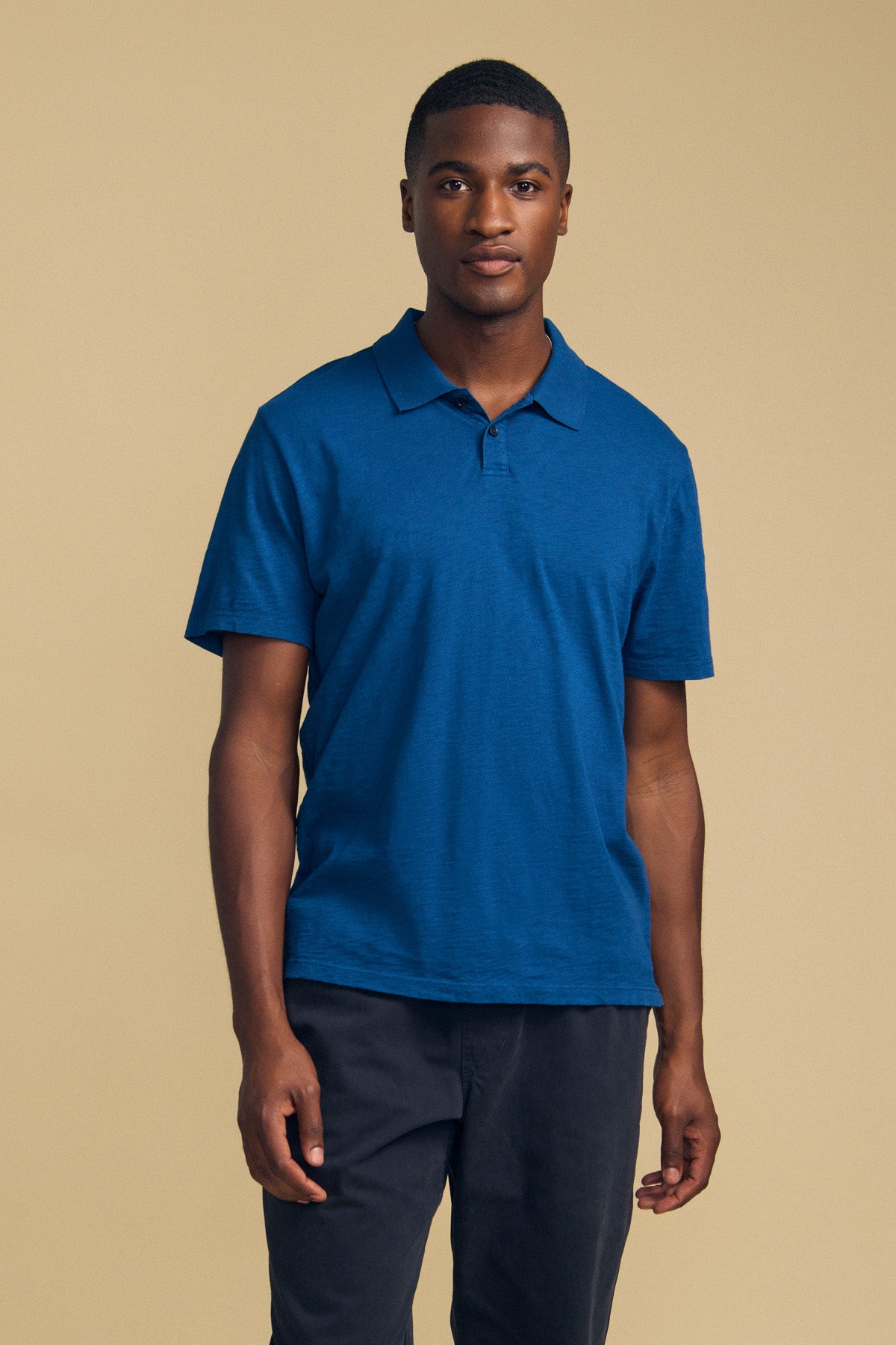 A man stands against a beige background, wearing the MORRIS POLO by Velvet by Graham & Spencer—a blue cotton slub polo with short sleeves and a ribbed collar—paired with dark pants. He looks directly at the camera with a neutral expression.-41859749150913