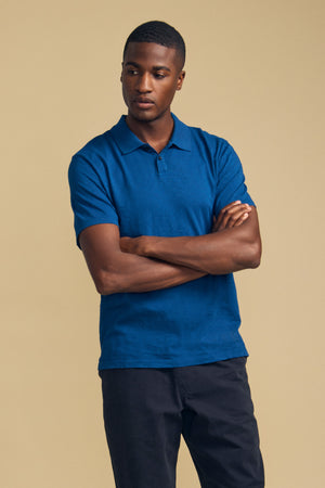 A man stands with arms crossed, wearing the MORRIS POLO by Velvet by Graham & Spencer—a blue cotton slub polo featuring short sleeves and a ribbed collar—paired with black pants against a plain beige background.