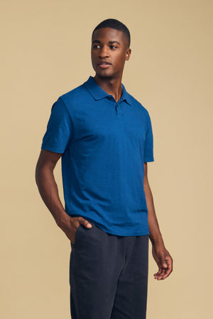 A man wearing the MORRIS POLO by Velvet by Graham & Spencer—a blue cotton slub polo with a ribbed collar and short sleeves—paired with black pants, stands against a plain beige background, looking slightly to his left with one hand in his pocket.
