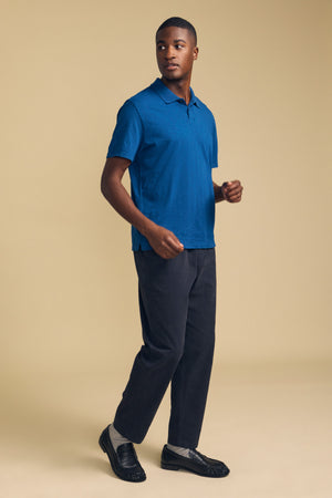 A man stands in a studio against a beige background wearing the MORRIS POLO by Velvet by Graham & Spencer—a blue cotton slub polo featuring short sleeves and a ribbed collar—paired with dark pants, gray socks, and black loafers.