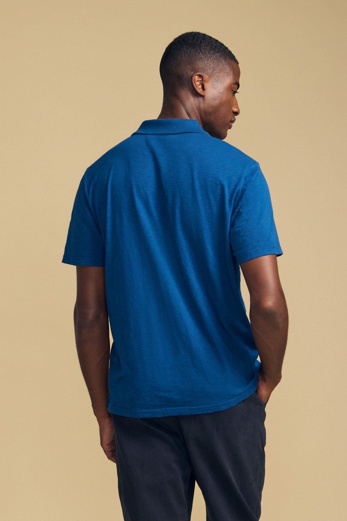 A man stands with his back to the camera, wearing the Velvet by Graham & Spencer MORRIS POLO in blue cotton slub, which features short sleeves and a ribbed collar, paired with dark pants against a beige background.-41859749380289
