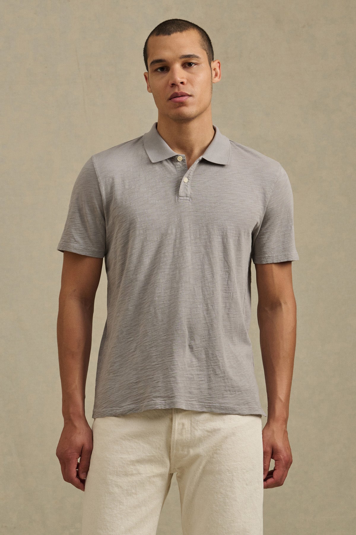 A man faces the camera against a plain beige background, wearing the MORRIS POLO by Velvet by Graham & Spencer in light gray with short sleeves, paired with off-white pants.-41338471055553