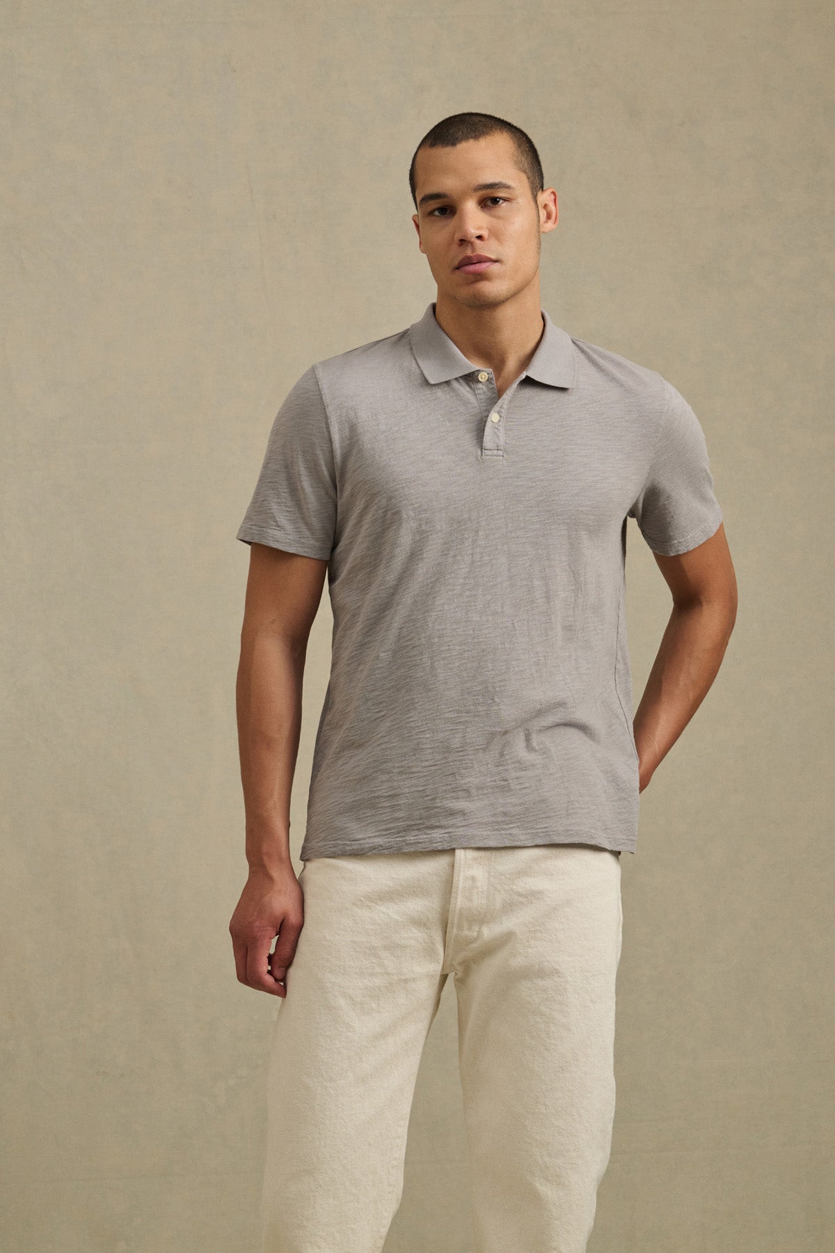 A man stands before a beige backdrop wearing the MORRIS POLO by Velvet by Graham & Spencer in light gray and off-white pants, with one hand behind his back.