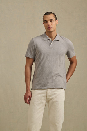 A man stands before a beige backdrop wearing the MORRIS POLO by Velvet by Graham & Spencer in light gray and off-white pants, with one hand behind his back.