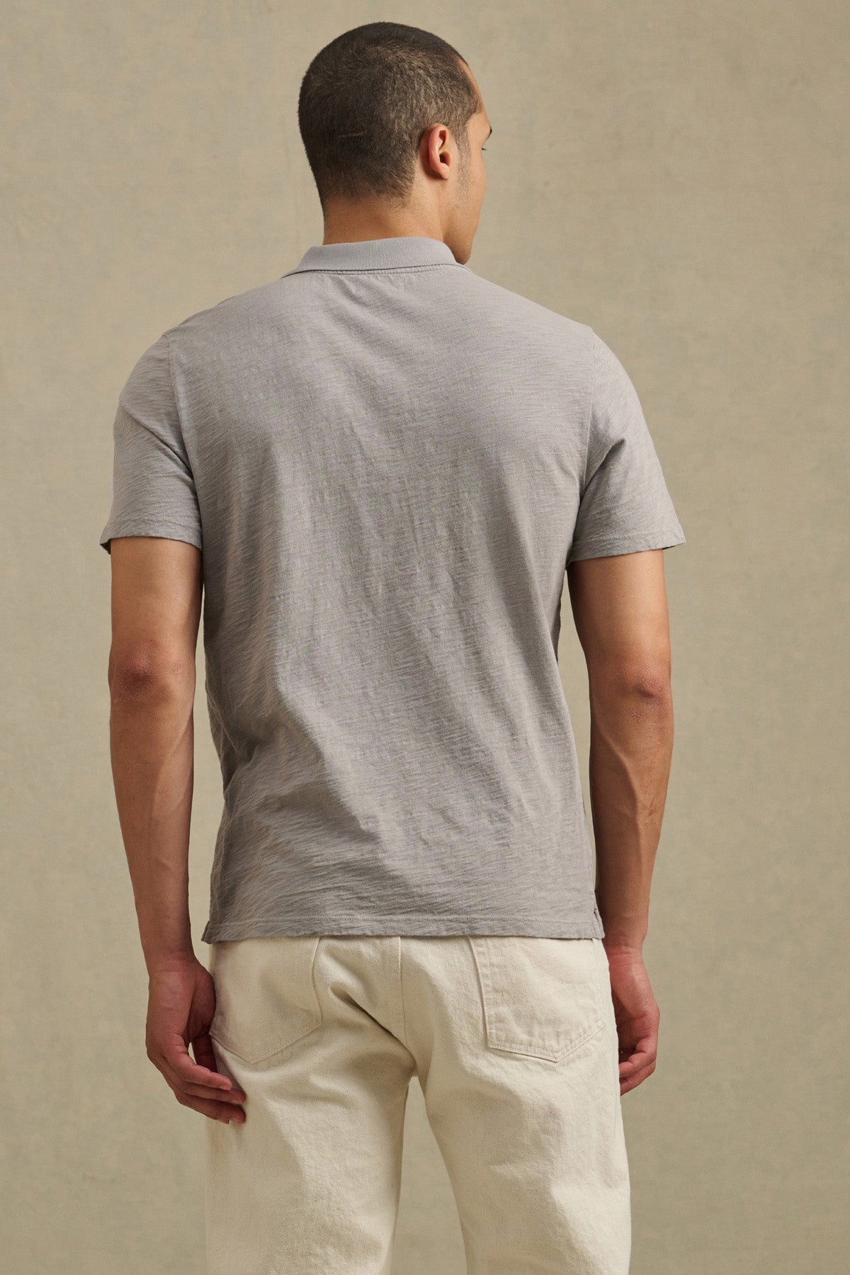 A man stands facing away, wearing the Velvet by Graham & Spencer MORRIS POLO in light gray and off-white pants against a plain beige background.