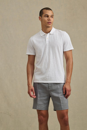 A man wears the MORRIS POLO by Velvet by Graham & Spencer, a white cotton slub short-sleeve polo, with gray shorts against a beige background.