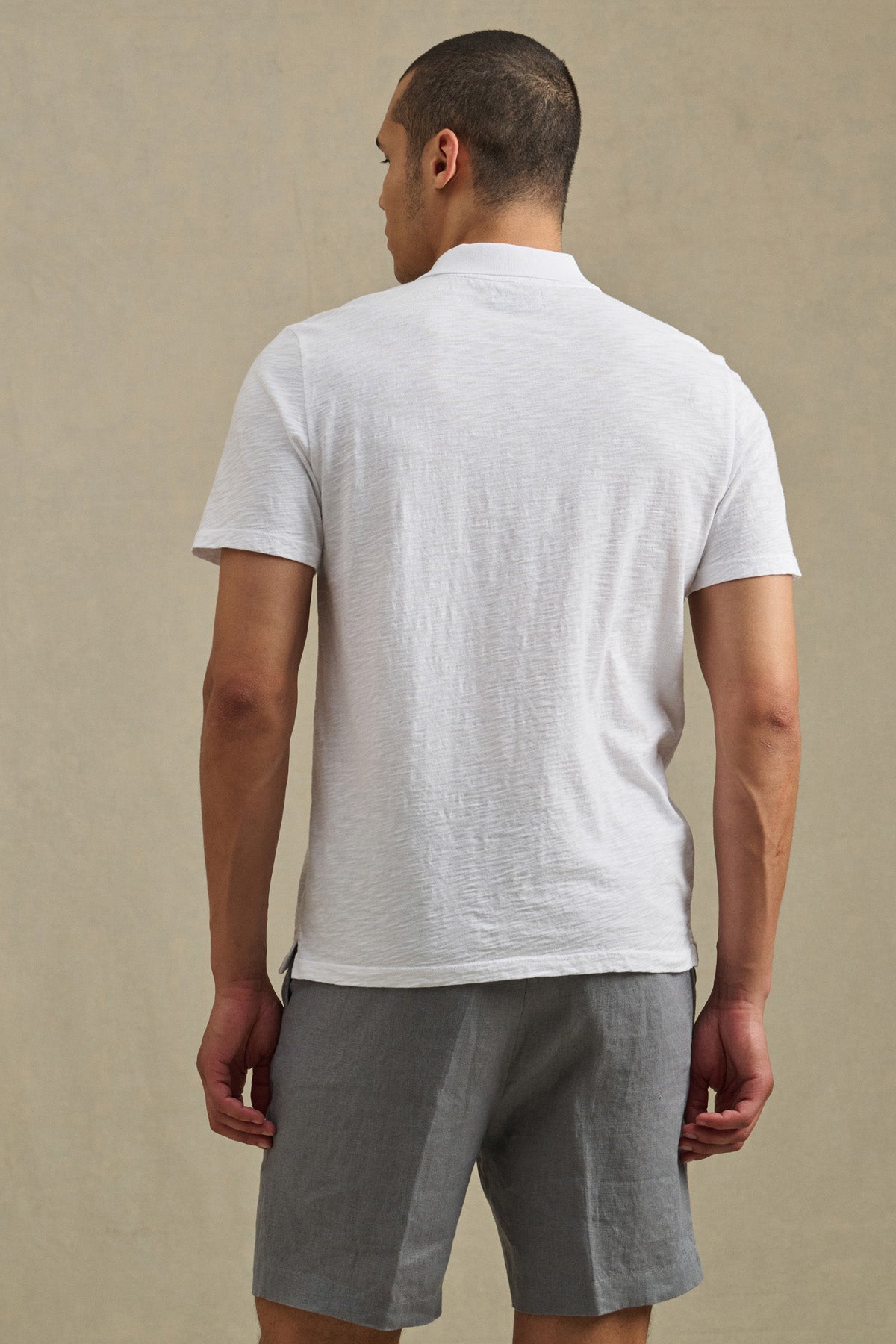 A man faces away, wearing the Velvet by Graham & Spencer MORRIS POLO in white with a two-button placket and gray shorts, set against a plain beige background.-41384130642113