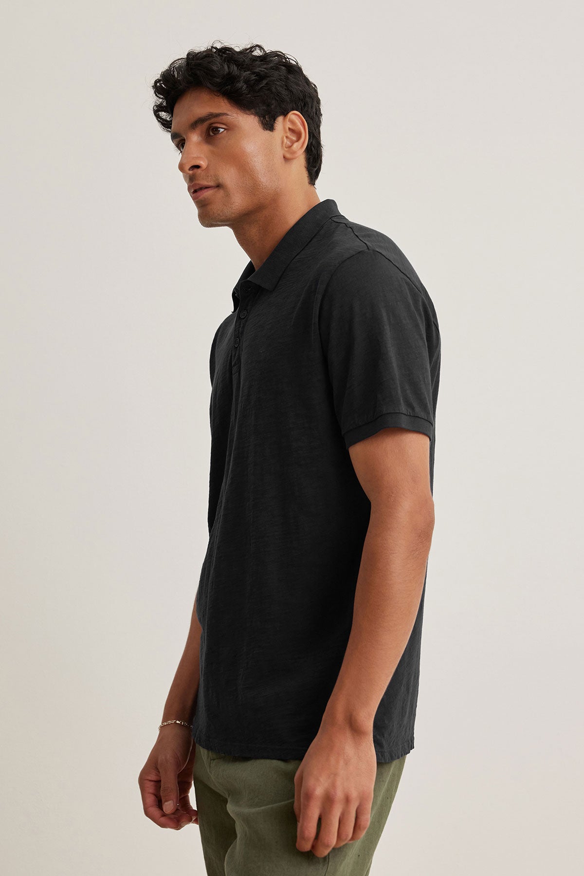   A man in a Velvet by Graham & Spencer NIKO POLO, a black short-sleeve shirt crafted from soft Peruvian cotton, paired with green pants, gazes sideways against a plain backdrop. 