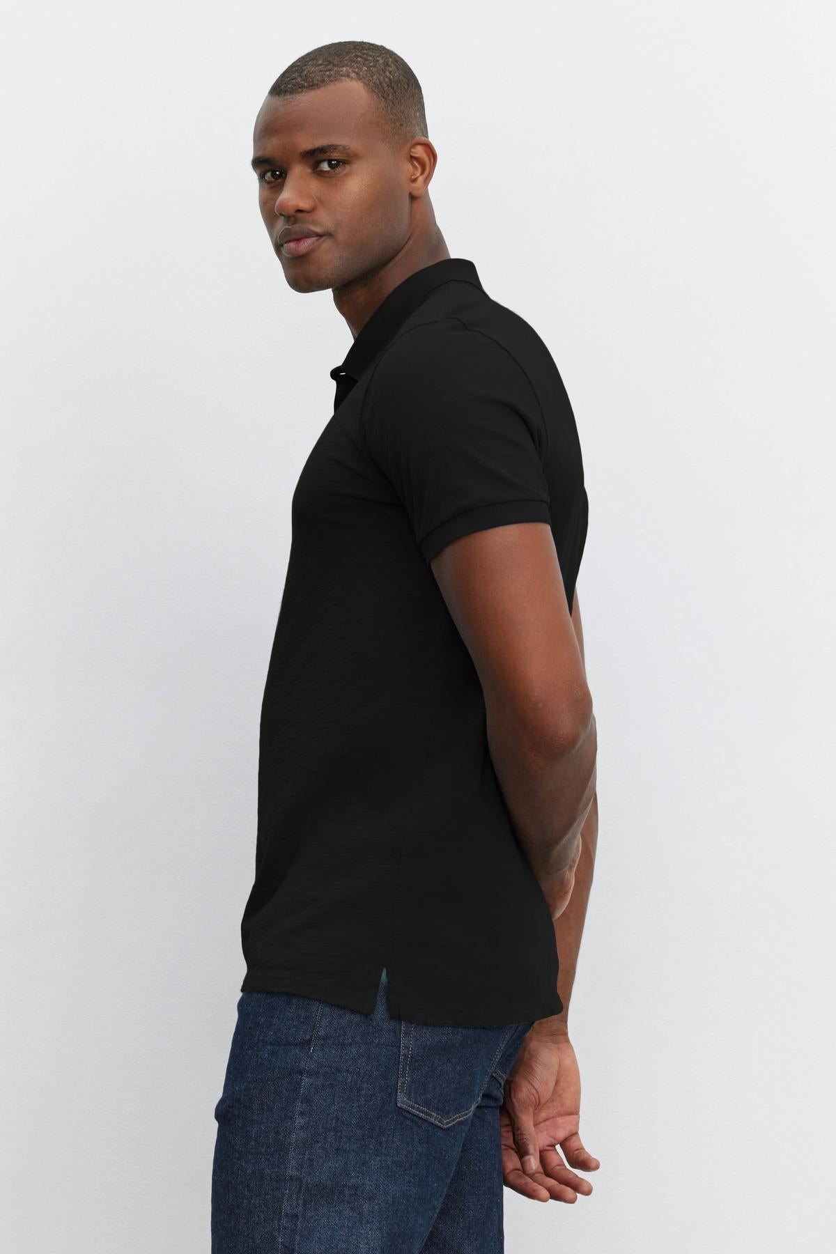 A young man in a black NIKO POLO polo shirt by Velvet by Graham & Spencer and jeans, standing sideways, looks over his shoulder.-36890752876737