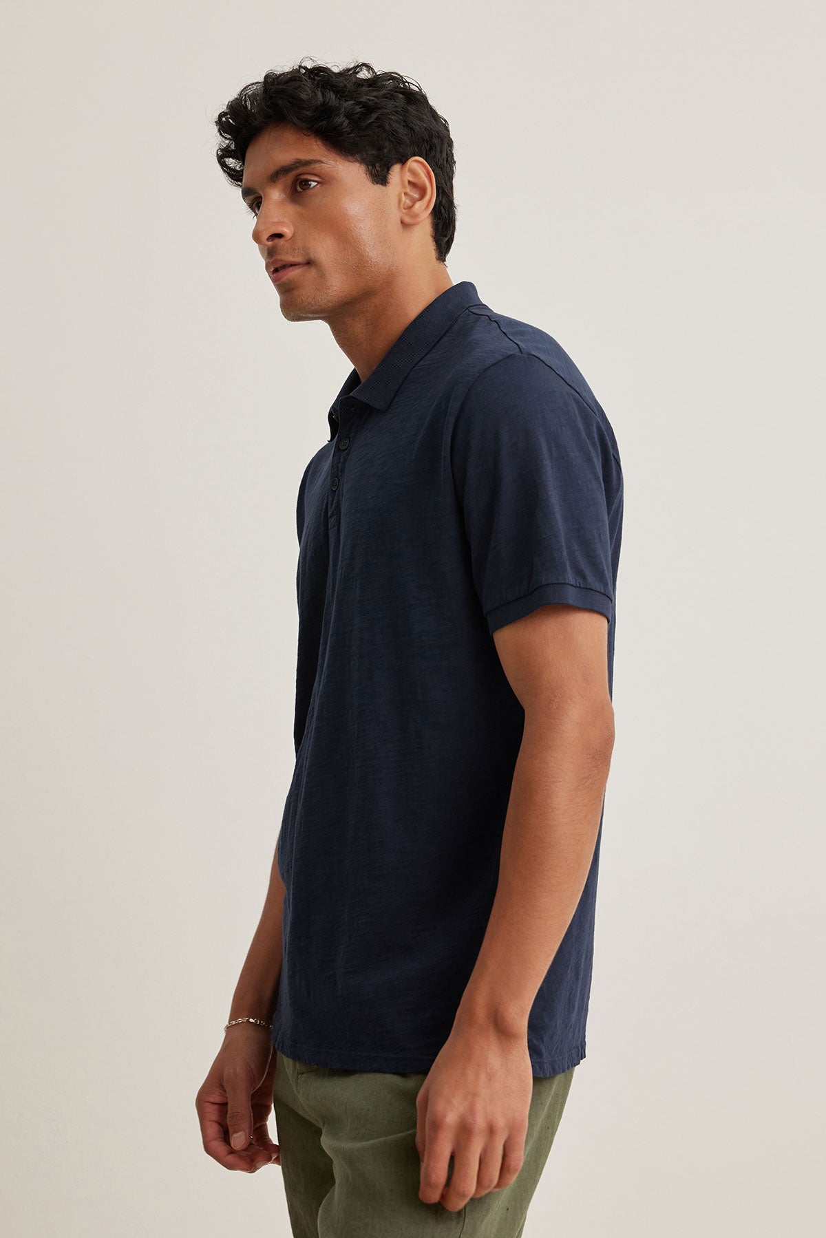   A man in a NIKO POLO from Velvet by Graham & Spencer, featuring a versatile navy blue color and 3-button placket, pairs it with green pants against a plain background. 