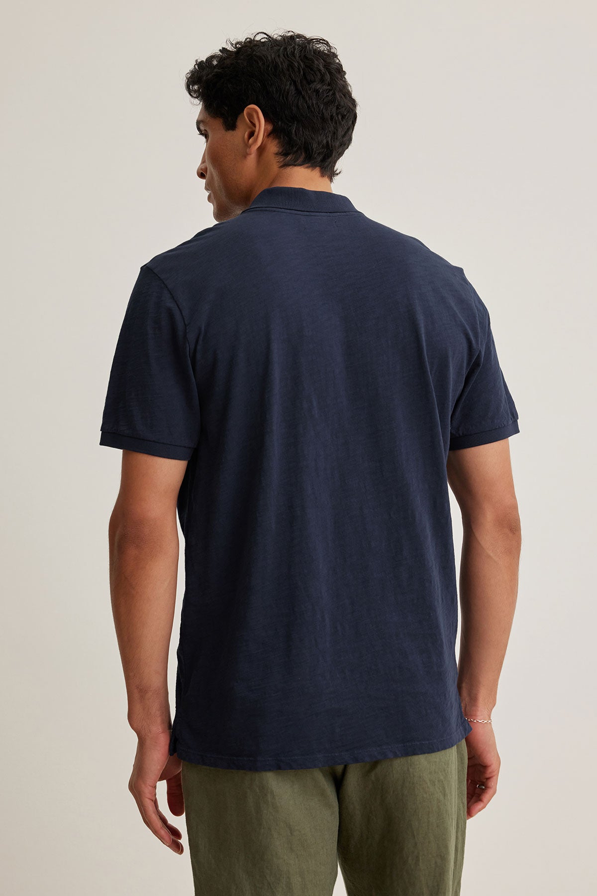 Someone in a Velvet by Graham & Spencer NIKO POLO, navy with 3 buttons, and olive pants stands facing away against a neutral background.-38800733700289