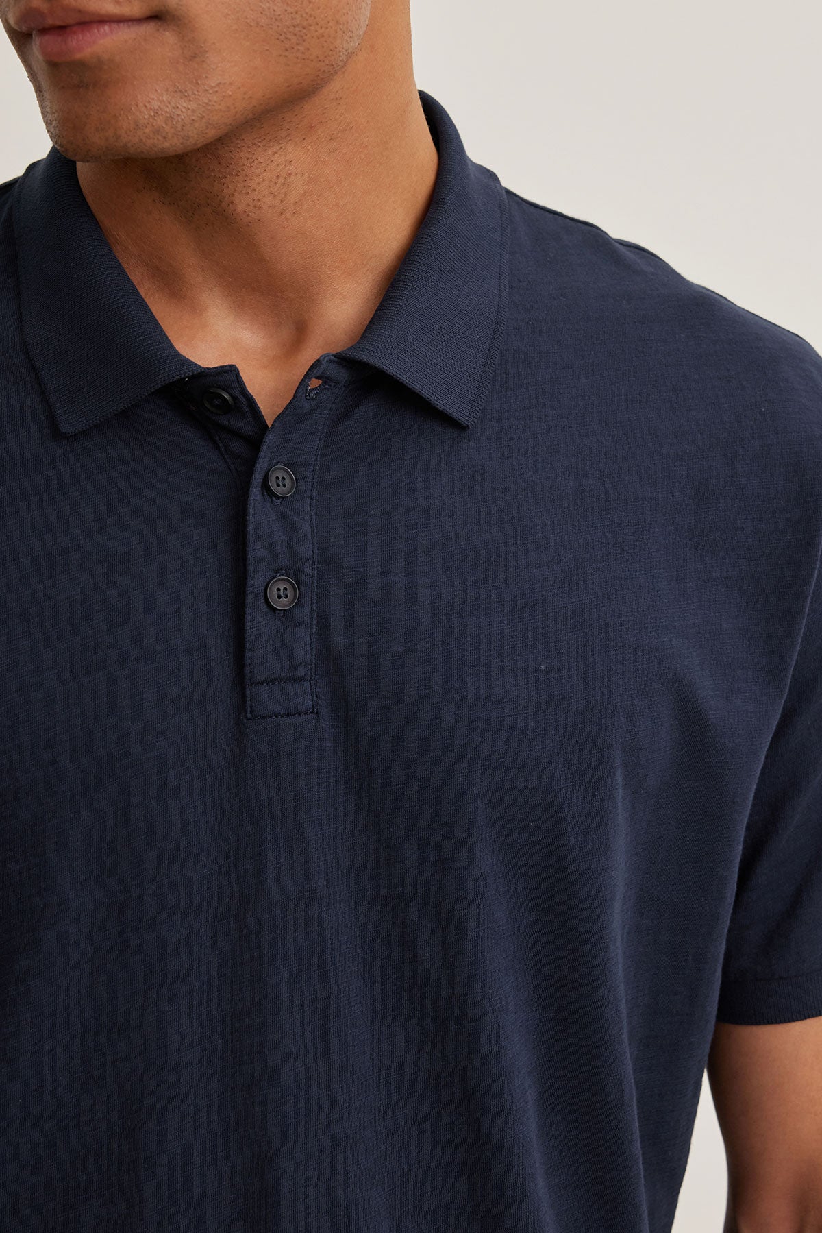   A man in the NIKO POLO by Velvet by Graham & Spencer, made of luxurious navy blue Peruvian cotton with a buttoned collar, stands against a neutral background. This shirt epitomizes versatile wear, effortlessly combining style and comfort for any occasion. 