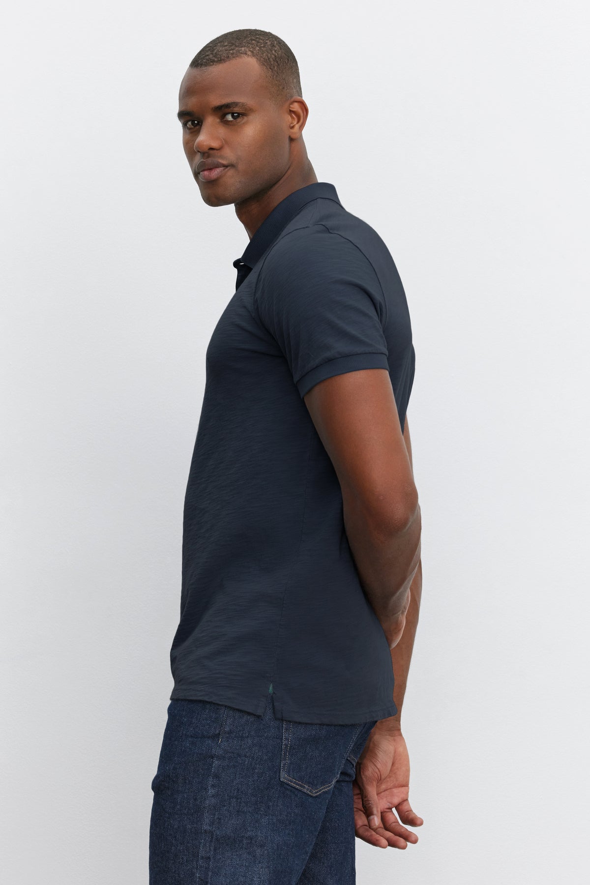 A man wearing a dark NIKO POLO by Velvet by Graham & Spencer shirt and jeans, standing sideways, looking over his shoulder with a neutral expression.-36890752778433
