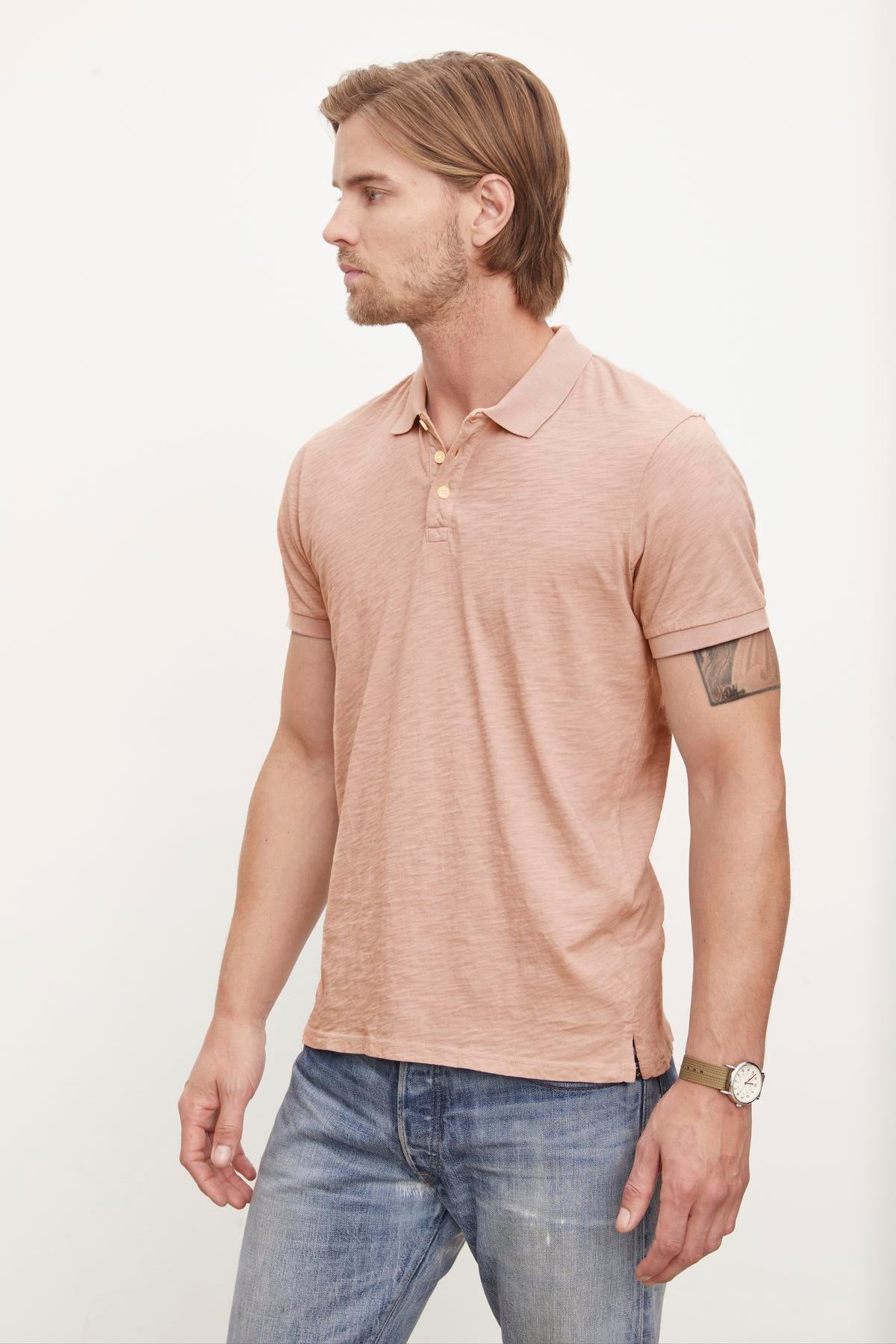 A man in a Velvet by Graham & Spencer NIKO POLO polo shirt with a heathered texture and blue jeans stands in profile, looking to the side, with a tattoo on his left arm and wearing a wristwatch.-36890746716353
