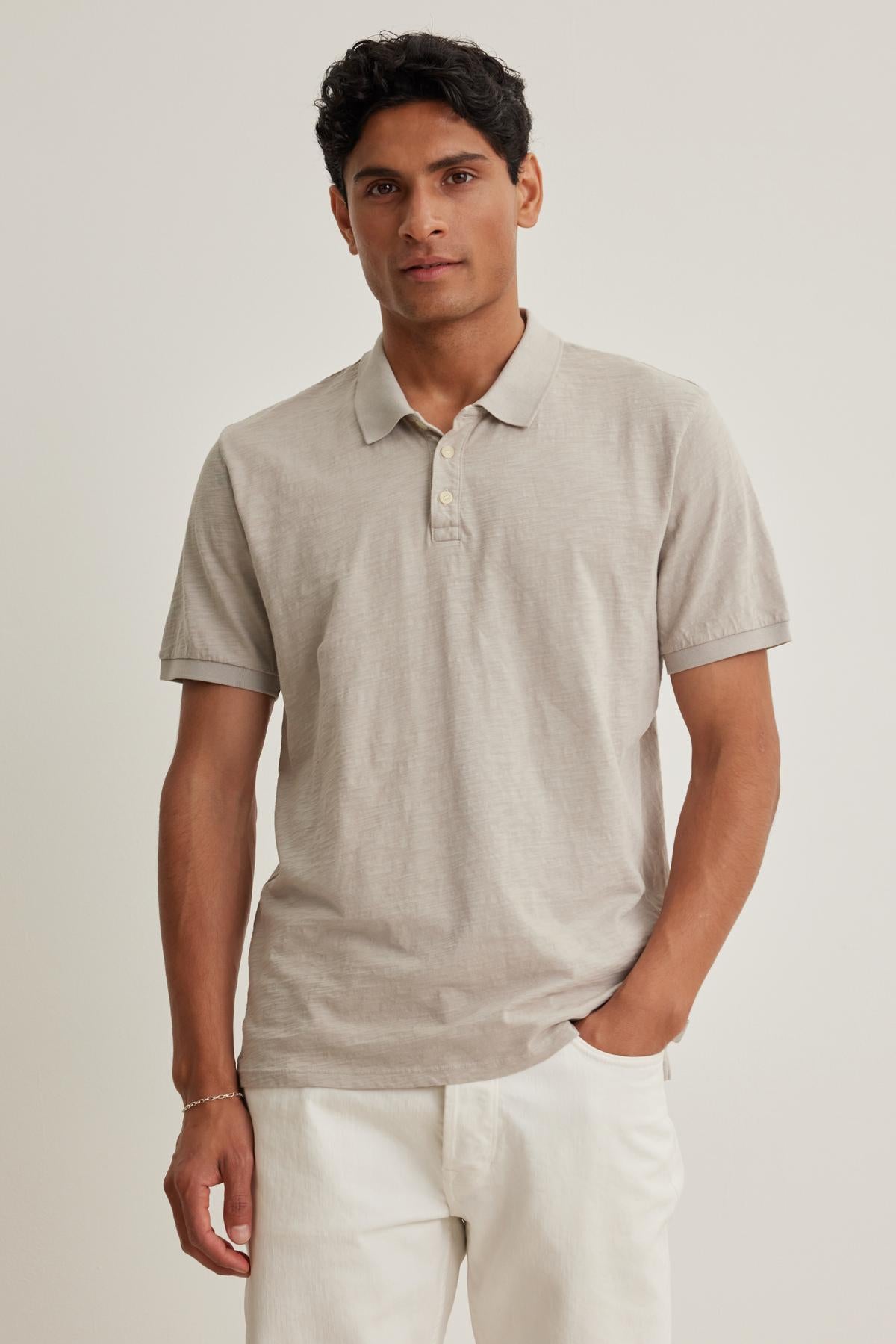   A person wears the versatile, lightweight NIKO POLO by Velvet by Graham & Spencer in beige, paired with white pants, standing against a plain background. 