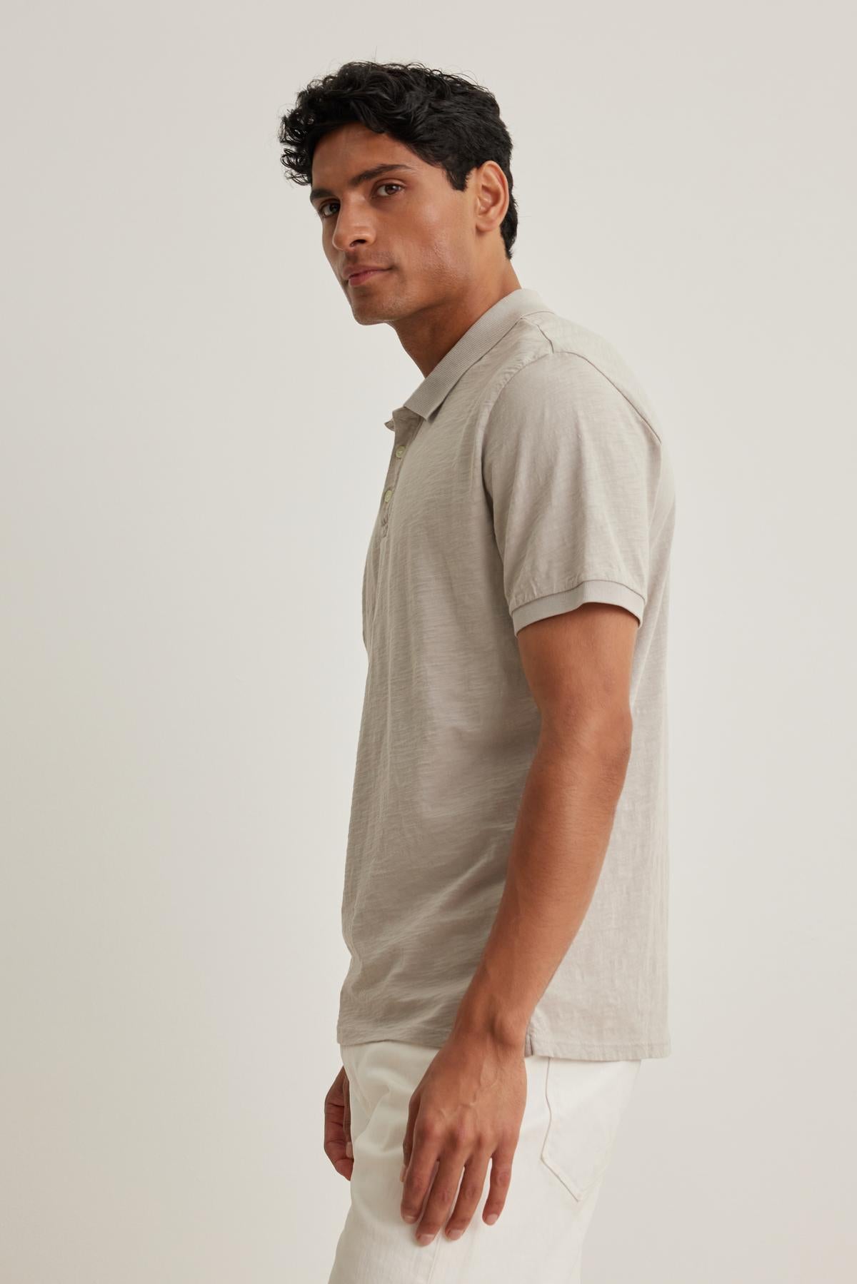   A man in a NIKO POLO by Velvet by Graham & Spencer and white pants stands sideways against a plain backdrop, showcasing the versatile wear that fuses comfort with style. 