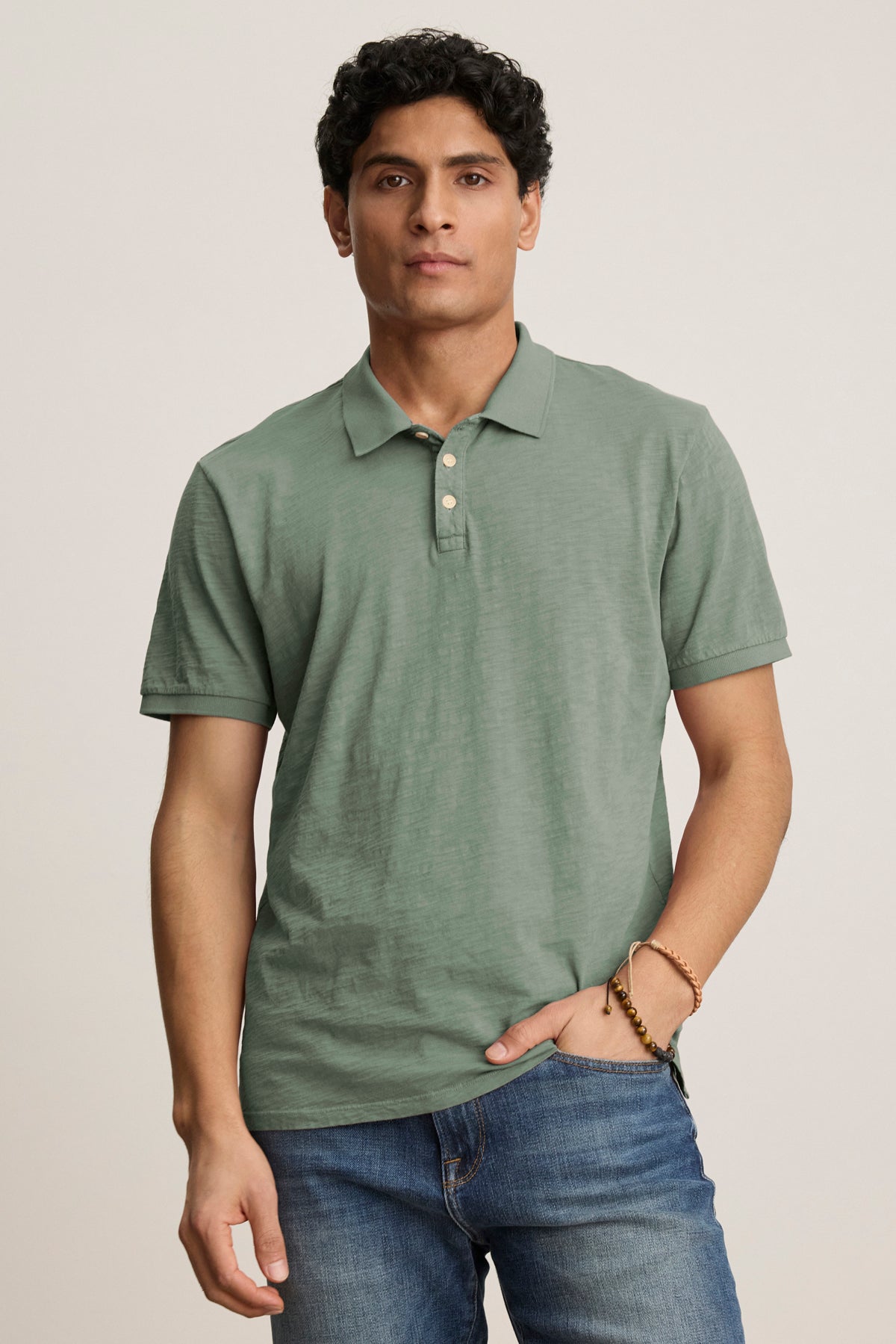   A man wears the Velvet by Graham & Spencer NIKO POLO in sage green, featuring a 3-button placket, paired with blue jeans, standing against a plain light background. 