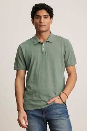 A man wears the Velvet by Graham & Spencer NIKO POLO in sage green, featuring a 3-button placket, paired with blue jeans, standing against a plain light background.