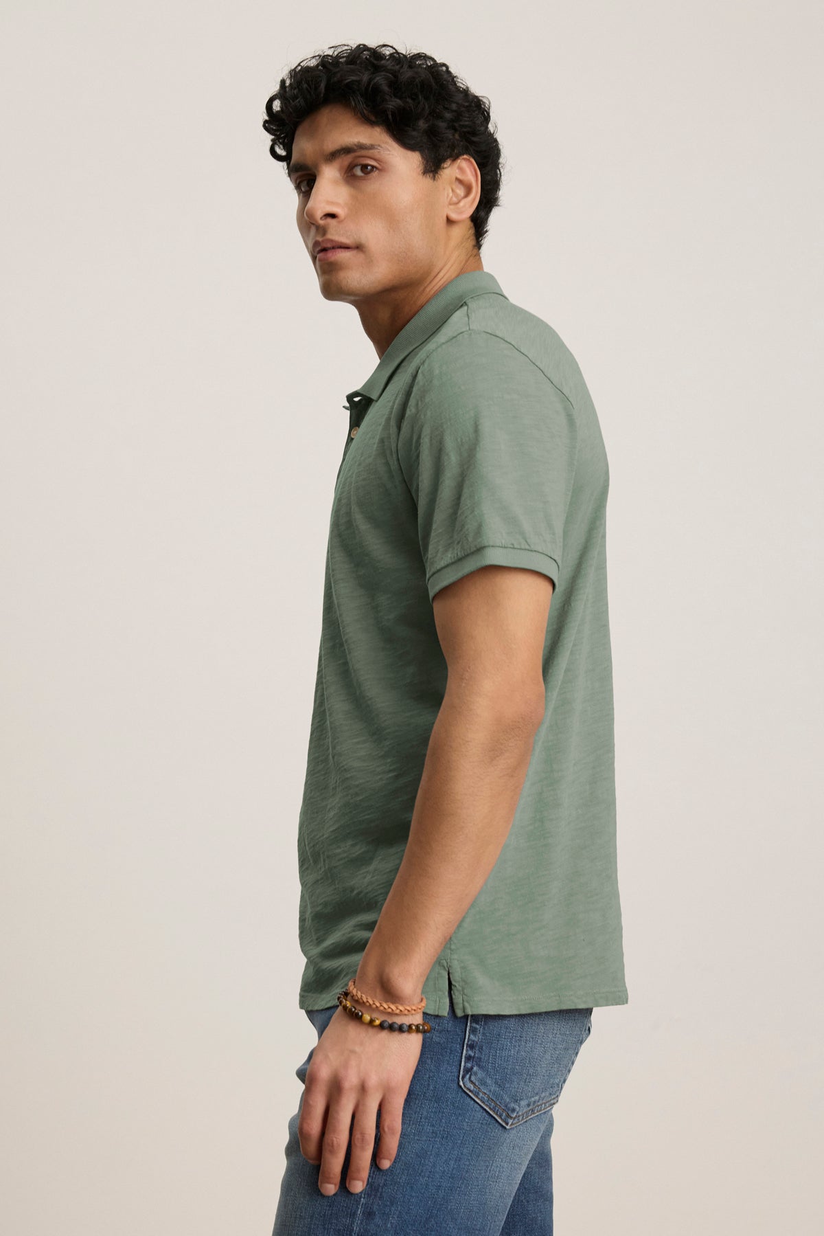   A person wearing the NIKO POLO by Velvet by Graham & Spencer—green, short-sleeve, 3-button placket—and blue jeans stands sideways against a plain light background. 