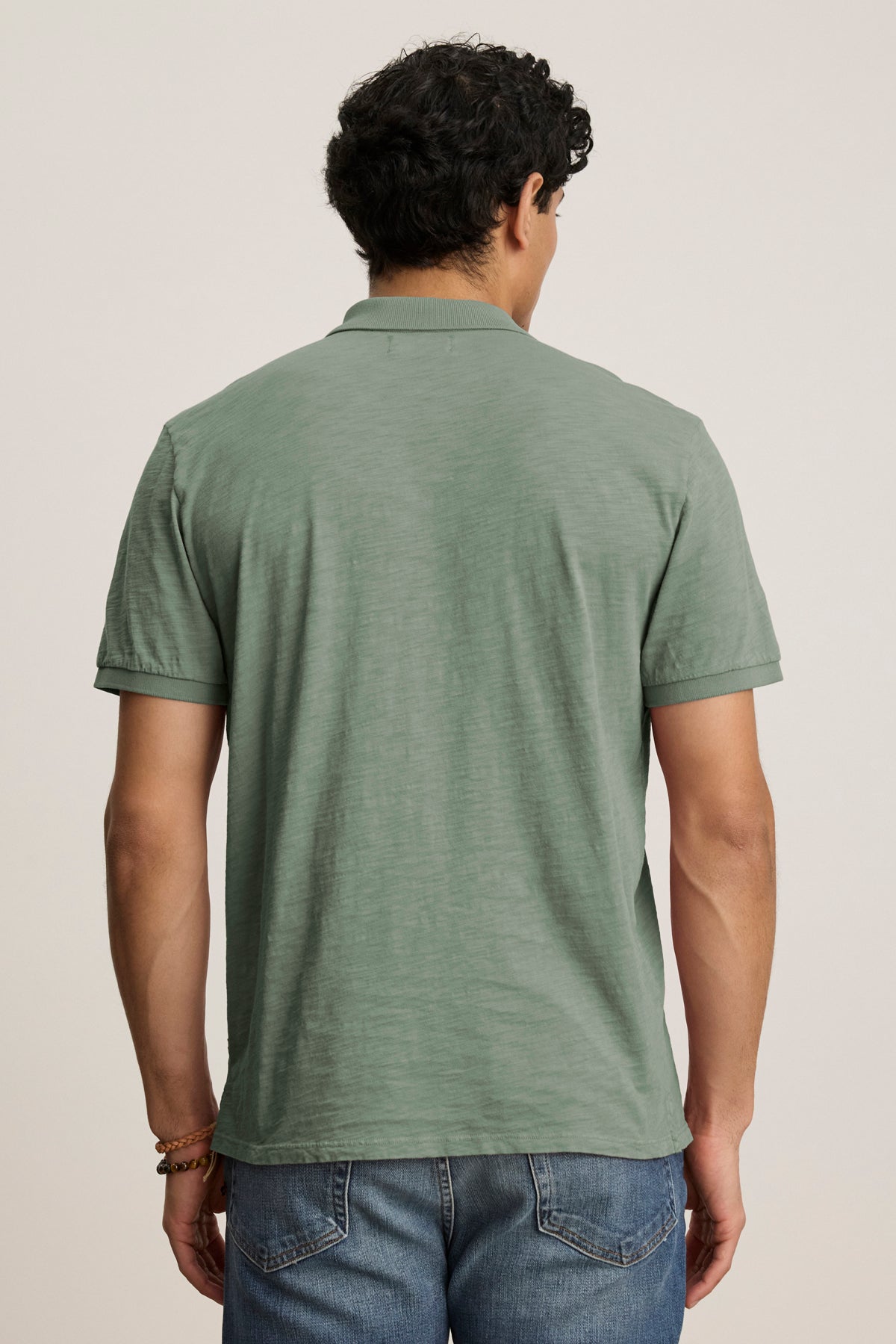   A man faces away from the camera, wearing the NIKO POLO by Velvet by Graham & Spencer—a light green Peruvian cotton polo with a 3-button placket—paired with blue jeans, set against a plain background. 