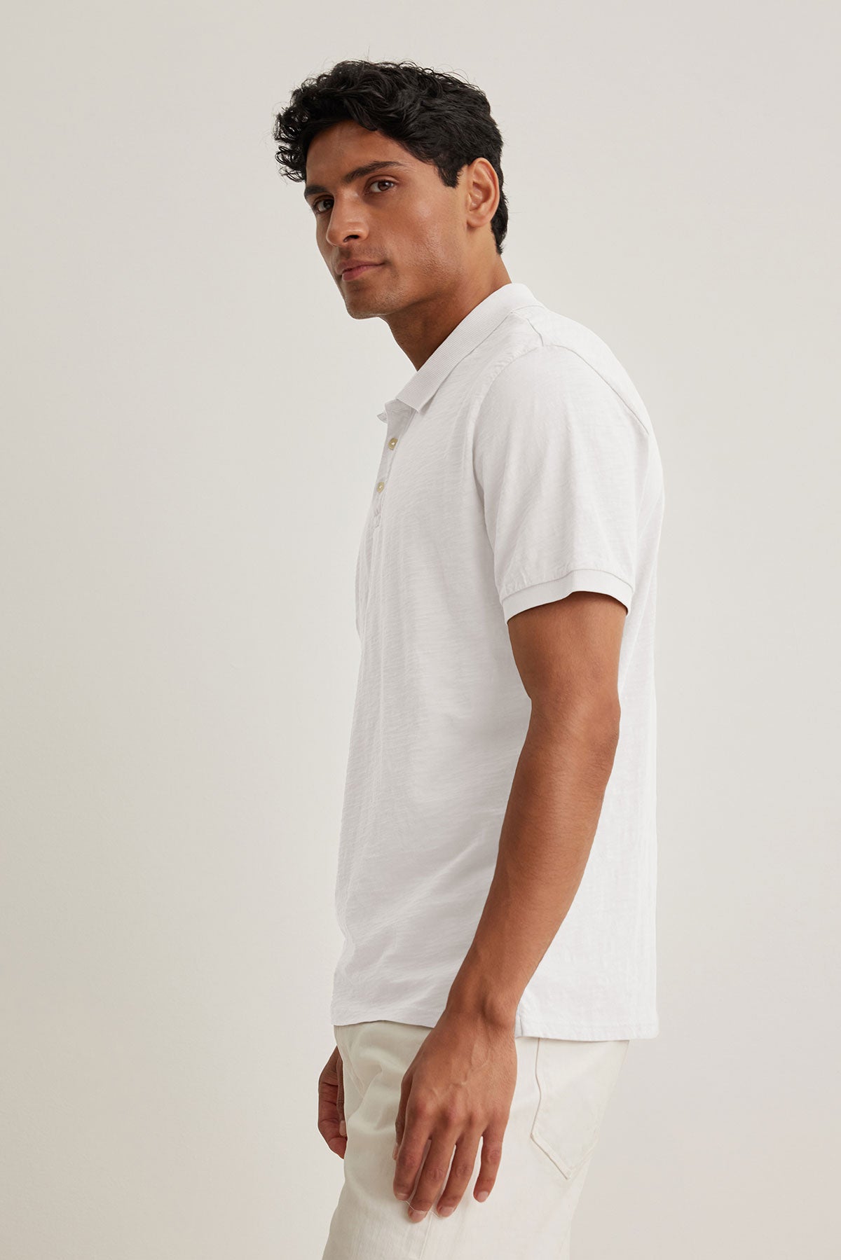   A man wearing the NIKO POLO by Velvet by Graham & Spencer, made from lightweight cotton, and beige pants stands sideways against a plain background, highlighting the versatility of this ensemble. 