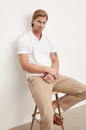 A man sitting on a stool wearing a Velvet by Graham & Spencer NIKO POLO in white cotton slub fabric and tan pants.