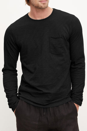 A person wears the SIMEON TEE—a long-sleeve black Peruvian cotton slub crew-neck shirt with a chest pocket by Velvet by Graham & Spencer—paired with dark pants, standing hands-in-pockets against a plain background.