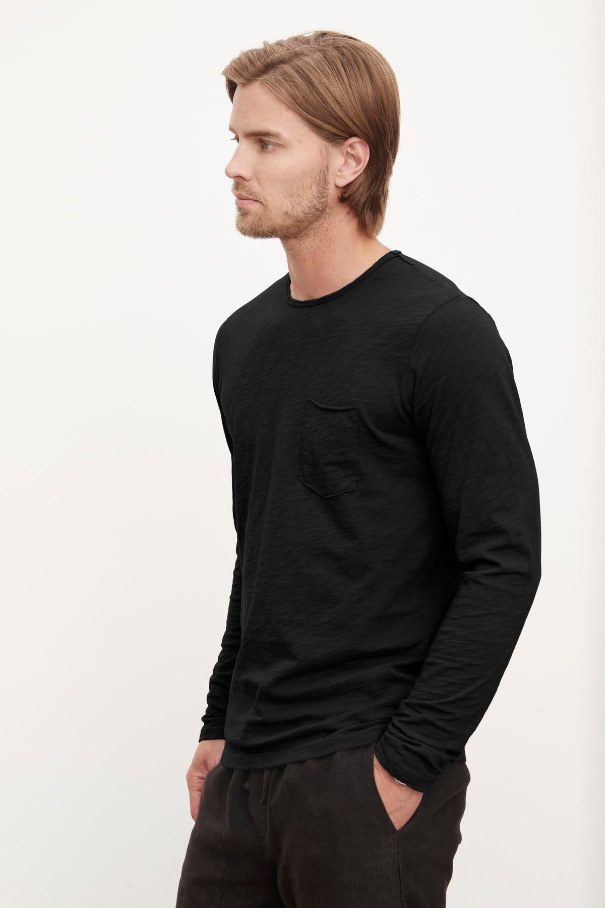 Man wearing a black long-sleeve SIMEON RAW EDGE COTTON SLUB TEE with a chest pocket, standing profile from Velvet by Graham & Spencer.-36418291105985