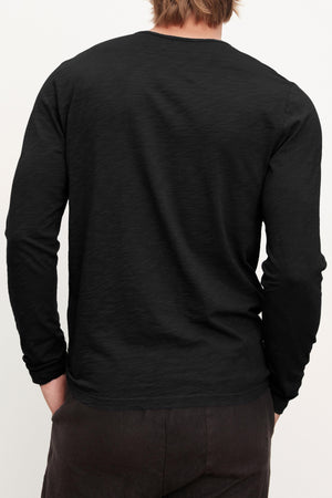 A person with light brown hair, shown from the back with hands in their pockets, wears the SIMEON TEE by Velvet by Graham & Spencer—a long-sleeve black shirt made from soft Peruvian cotton slub—paired with dark pants.