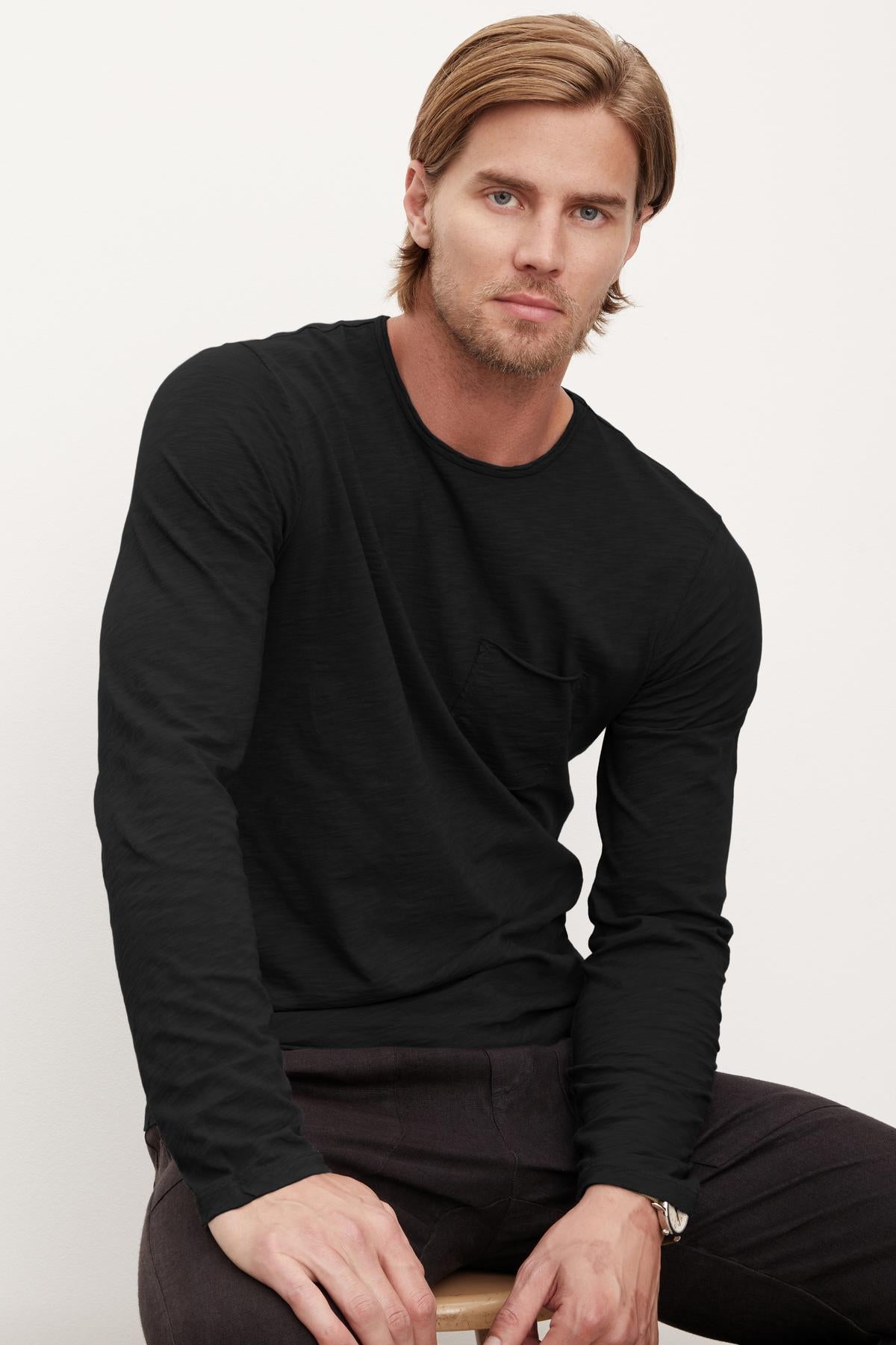   A man with medium-length hair wearing a black long-sleeve SIMEON RAW EDGE COTTON SLUB TEE with a pocket, posing for a portrait by Velvet by Graham & Spencer. 