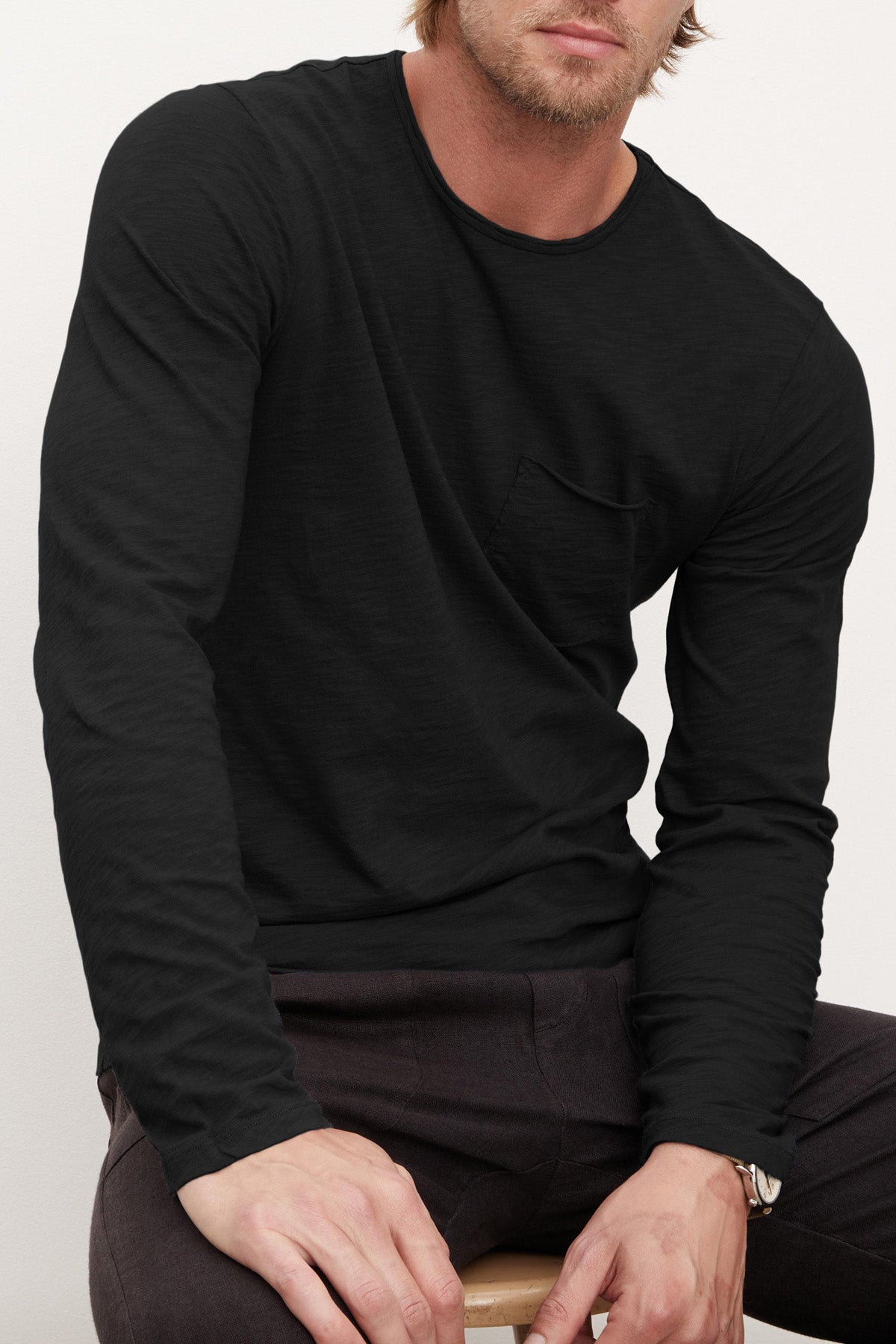 A man wearing the SIMEON TEE by Velvet by Graham & Spencer and dark pants sits on a wooden stool against a plain light background.