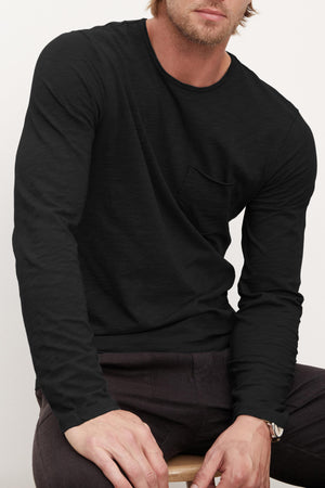 A man wearing the SIMEON TEE by Velvet by Graham & Spencer and dark pants sits on a wooden stool against a plain light background.