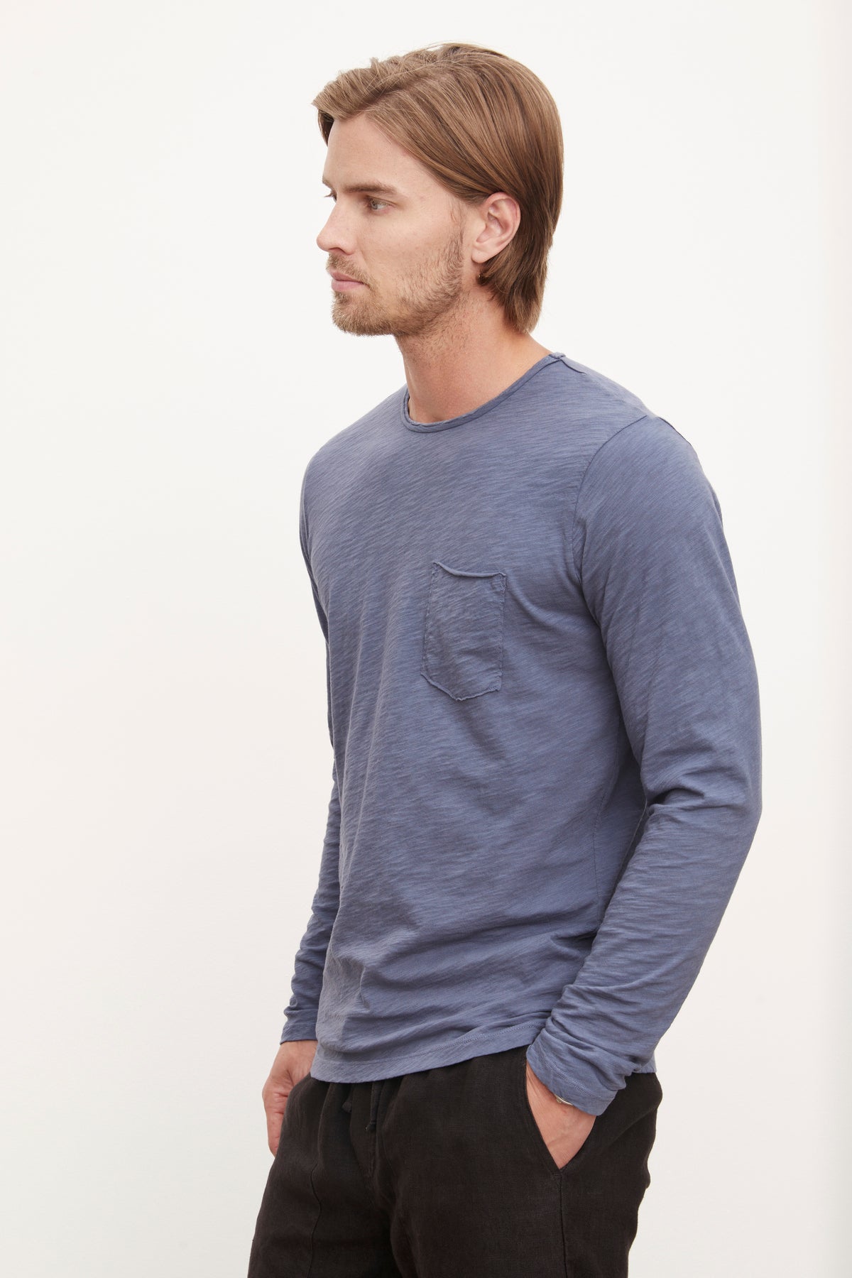  A man wearing a blue long sleeve SIMEON RAW EDGE COTTON SLUB TEE, made from soft slub cotton fabric and featuring raw-edge details on the sleeves by Velvet by Graham & Spencer. 
