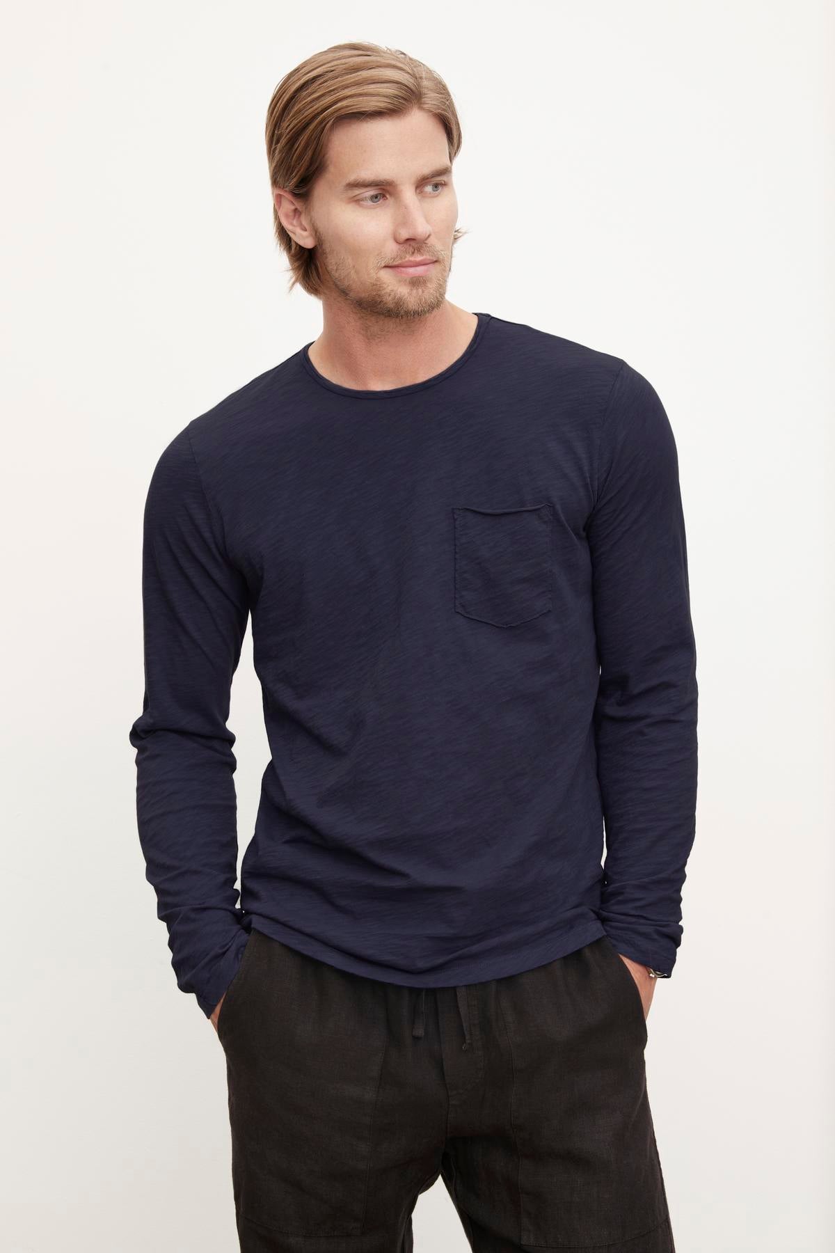   A man with neatly styled hair wearing a long-sleeve navy blue SIMEON RAW EDGE COTTON SLUB TEE with a pocket by Velvet by Graham & Spencer, posing with one hand in his pocket. 