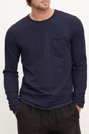 A man wears the SIMEON TEE by Velvet by Graham & Spencer—a long-sleeve navy crew neck made from Peruvian cotton slub with raw-edge details and a chest pocket—paired with dark pants, against a plain background.