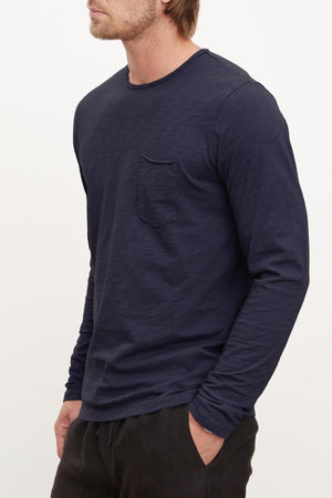 A man wearing the SIMEON TEE by Velvet by Graham & Spencer—a navy Peruvian cotton slub long-sleeve shirt with a chest pocket—paired with black pants, stands against a plain background.