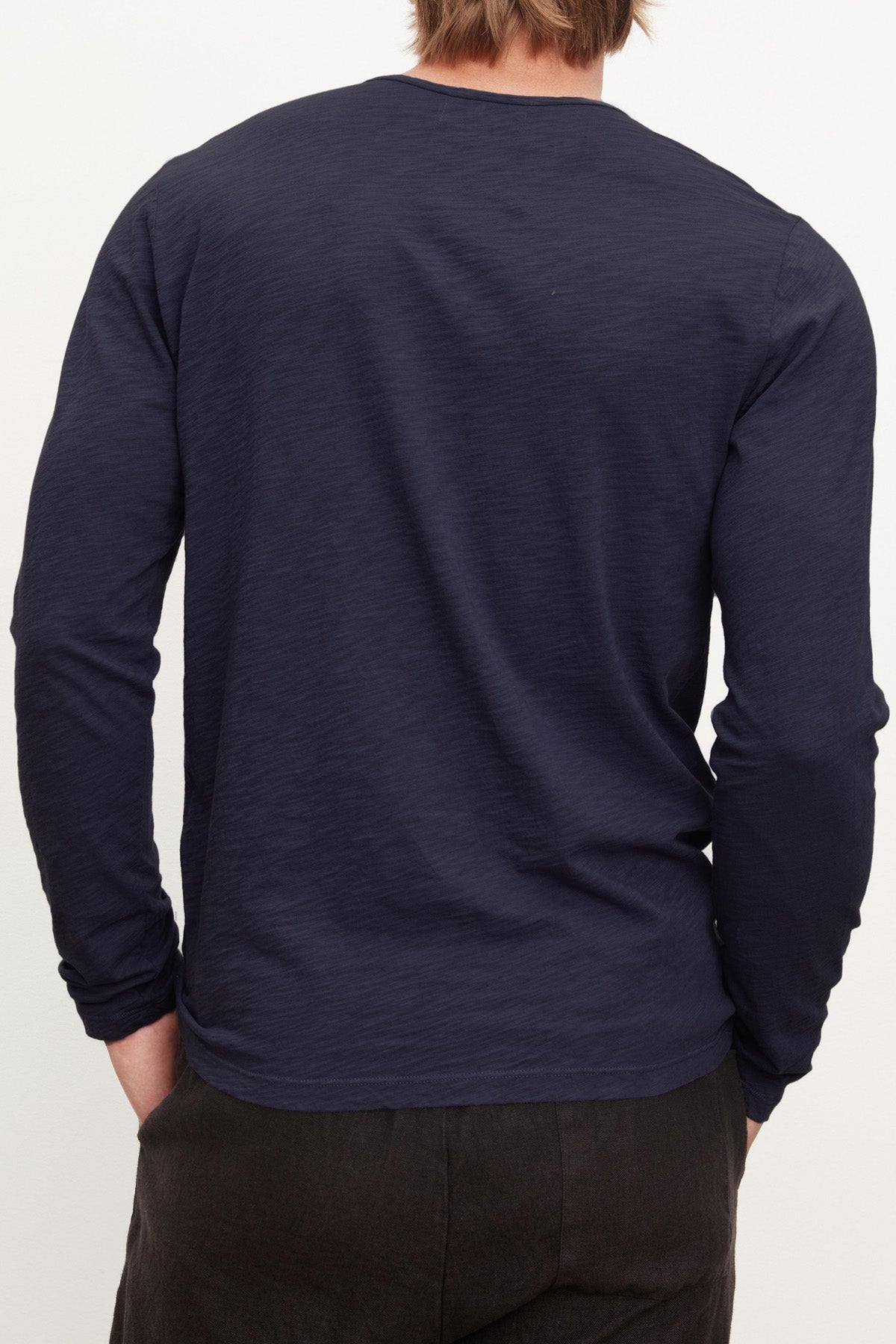 A person with light brown hair is seen from behind wearing a long-sleeved navy blue SIMEON TEE by Velvet by Graham & Spencer and dark pants, hands tucked into their pockets.