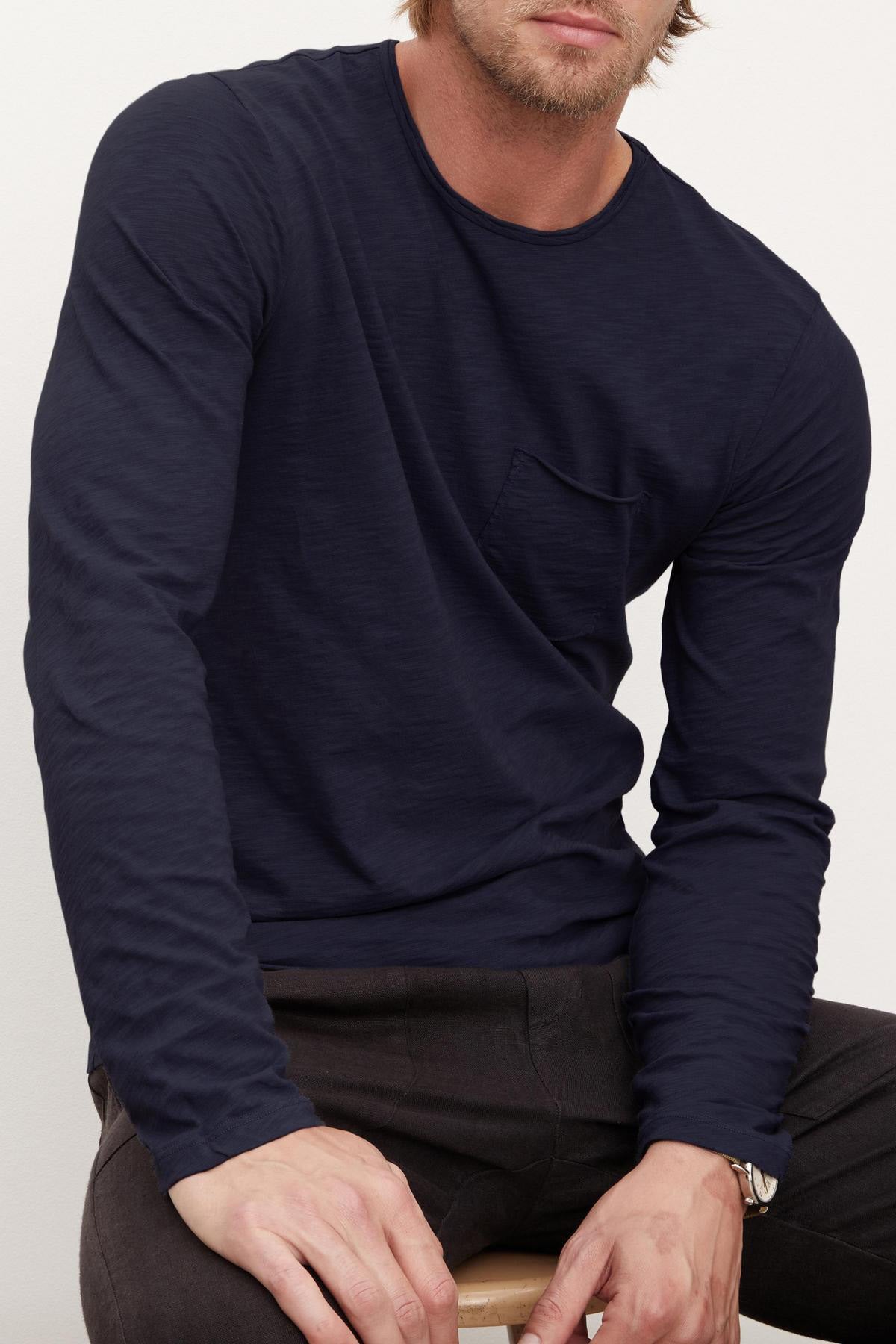A man sits on a stool wearing the SIMEON TEE by Velvet by Graham & Spencer, a long-sleeve dark navy Peruvian cotton slub shirt with a chest pocket. His face is partially out of frame.-40658337333441