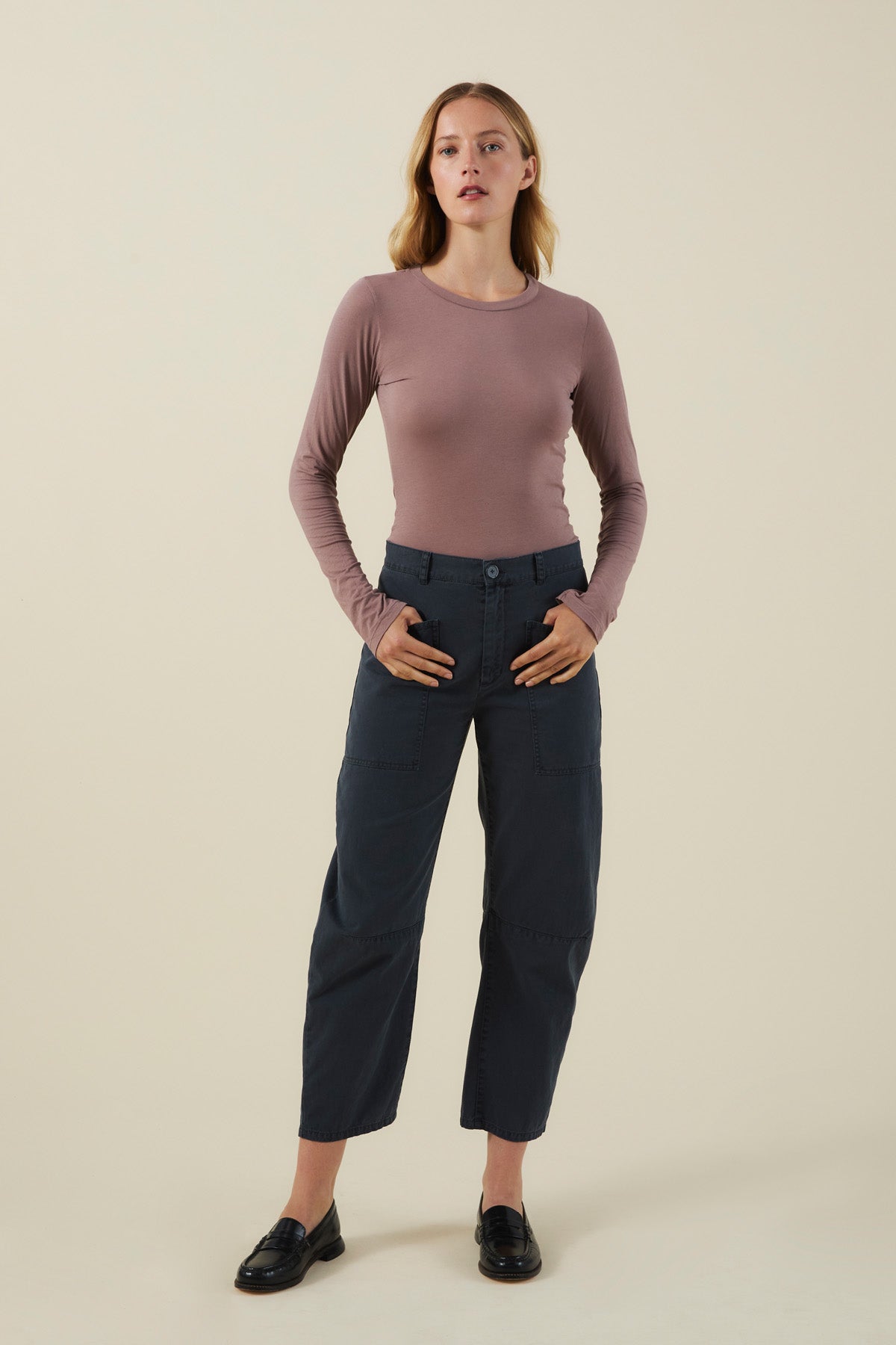 A model wears a long-sleeved dusty mauve shirt, dark cotton twill BRYLIE PANT with utilitarian patch pockets, and black loafers.