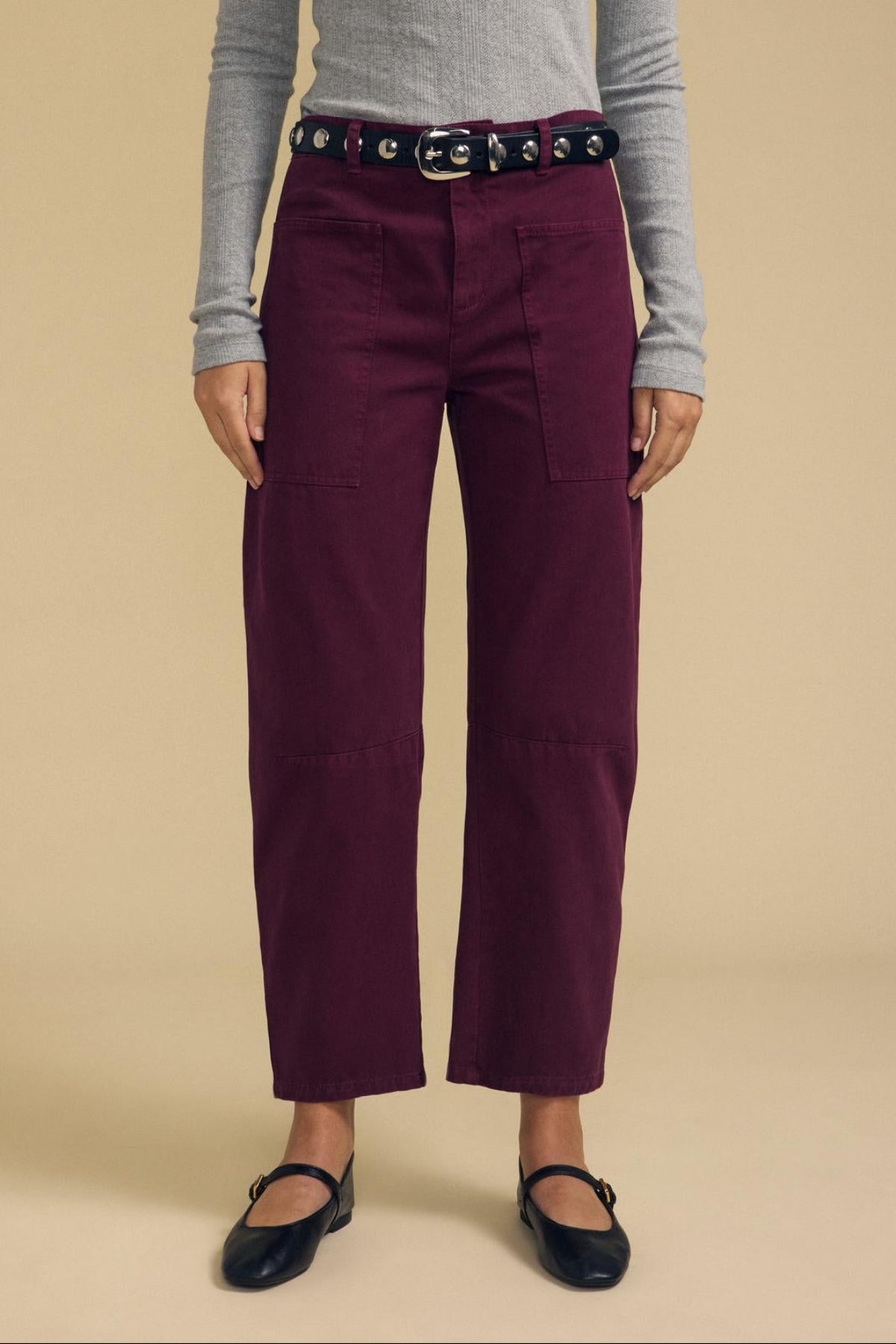 A person wearing a gray long-sleeve top, the BRYLIE PANT by Velvet by Graham & Spencer—structured burgundy cotton twill barrel leg trousers with utilitarian patch pockets—a black belt with grommets, and black ballet flats, standing against a beige background.