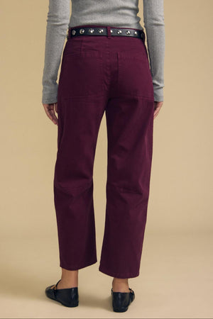 Clad in the BRYLIE PANT by Velvet by Graham & Spencer—a pair of structured cotton twill barrel leg trousers in burgundy—this individual pairs them with a gray long-sleeve shirt, black belt featuring silver grommets, and black flats, standing with their back to the camera against a beige background.