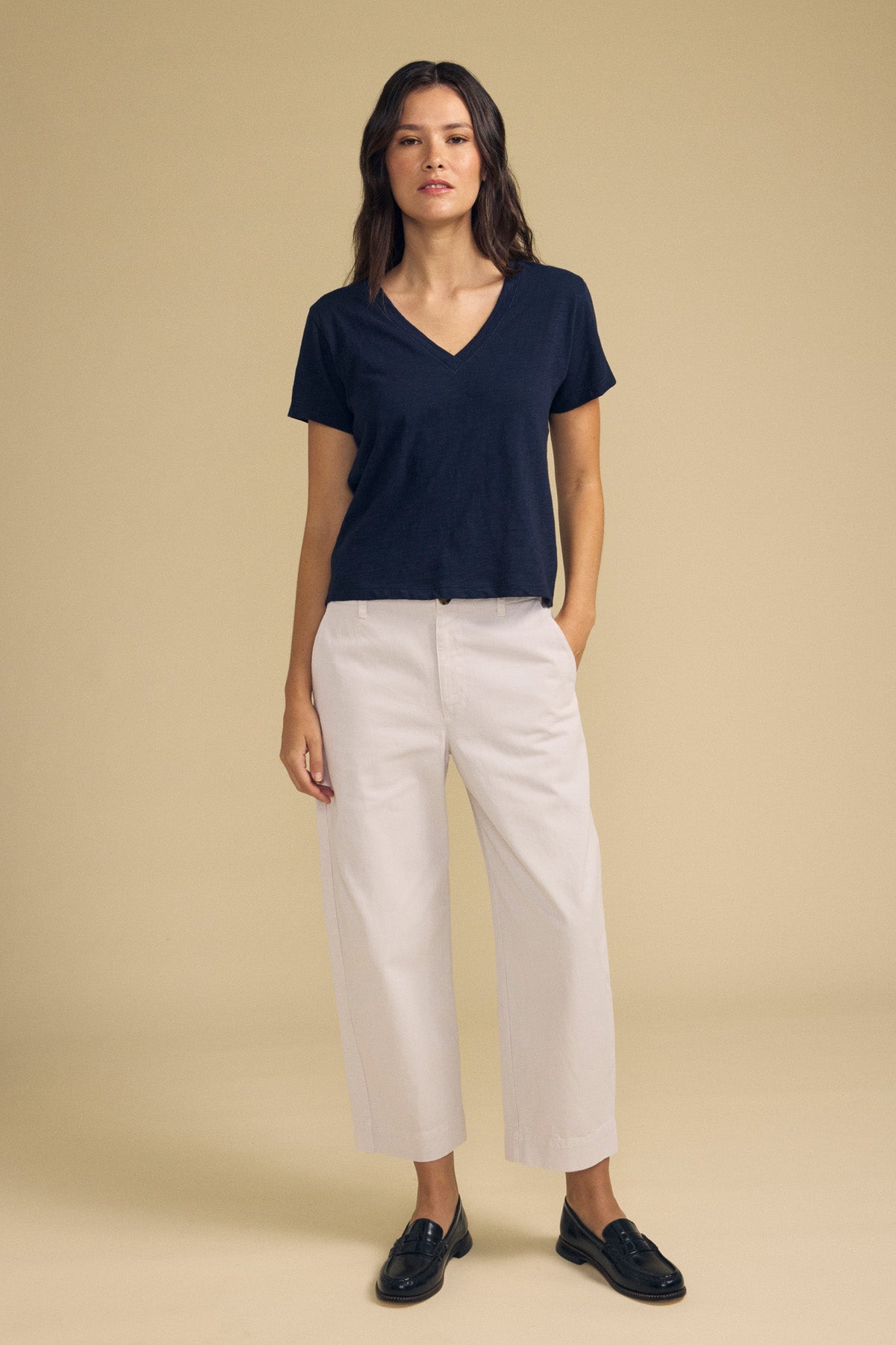 A woman stands against a beige background wearing a navy V-neck T-shirt, the DARIA PANT by Velvet by Graham & Spencer in white wide-leg style, and black loafers.-41814560374977