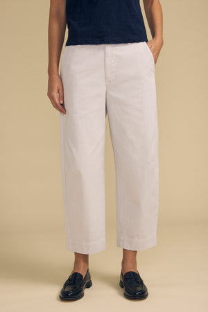 A person wearing a navy shirt, the DARIA PANT by Velvet by Graham & Spencer in light beige, and black loafers stands with one hand in their pocket against a plain beige background.