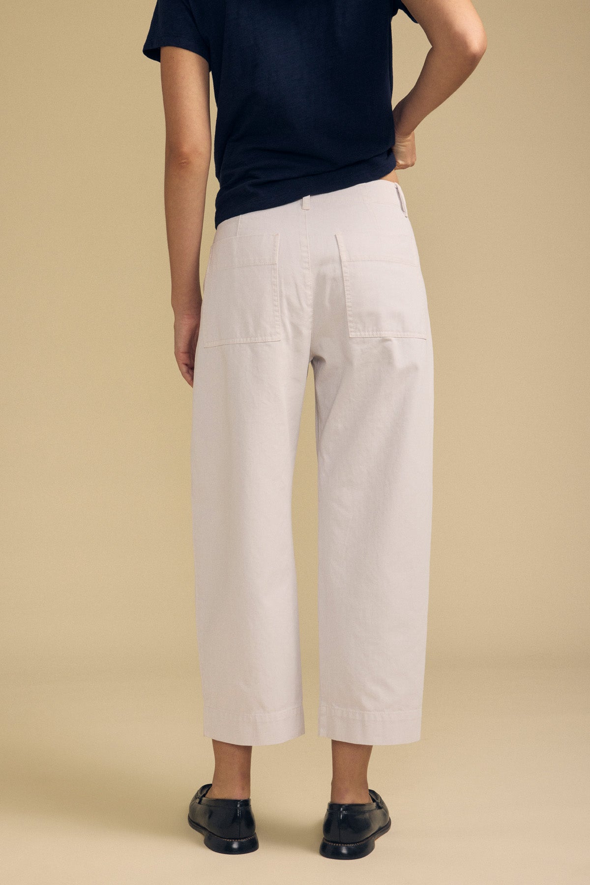 A person stands with their back to the camera against a beige background, wearing a black short-sleeve shirt, black shoes, and the DARIA PANT in white wide-leg cropped style by Velvet by Graham & Spencer.-41814560309441